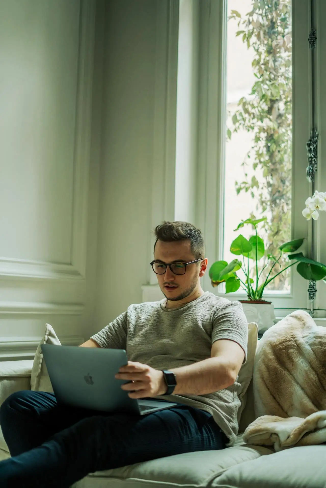 Homem sentado em um sofá de sala clara, usando um notebook em casa, com óculos e camiseta cinza. Ao fundo há janelas grandes, plantas verdes e flores brancas ao lado do vaso.