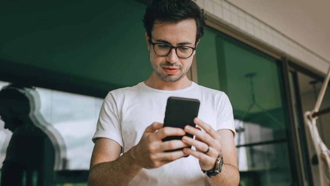 Homem usando smartphone em ambiente urbano, com óculos e camiseta branca, olhando para a tela enquanto segura o celular com as duas mãos.