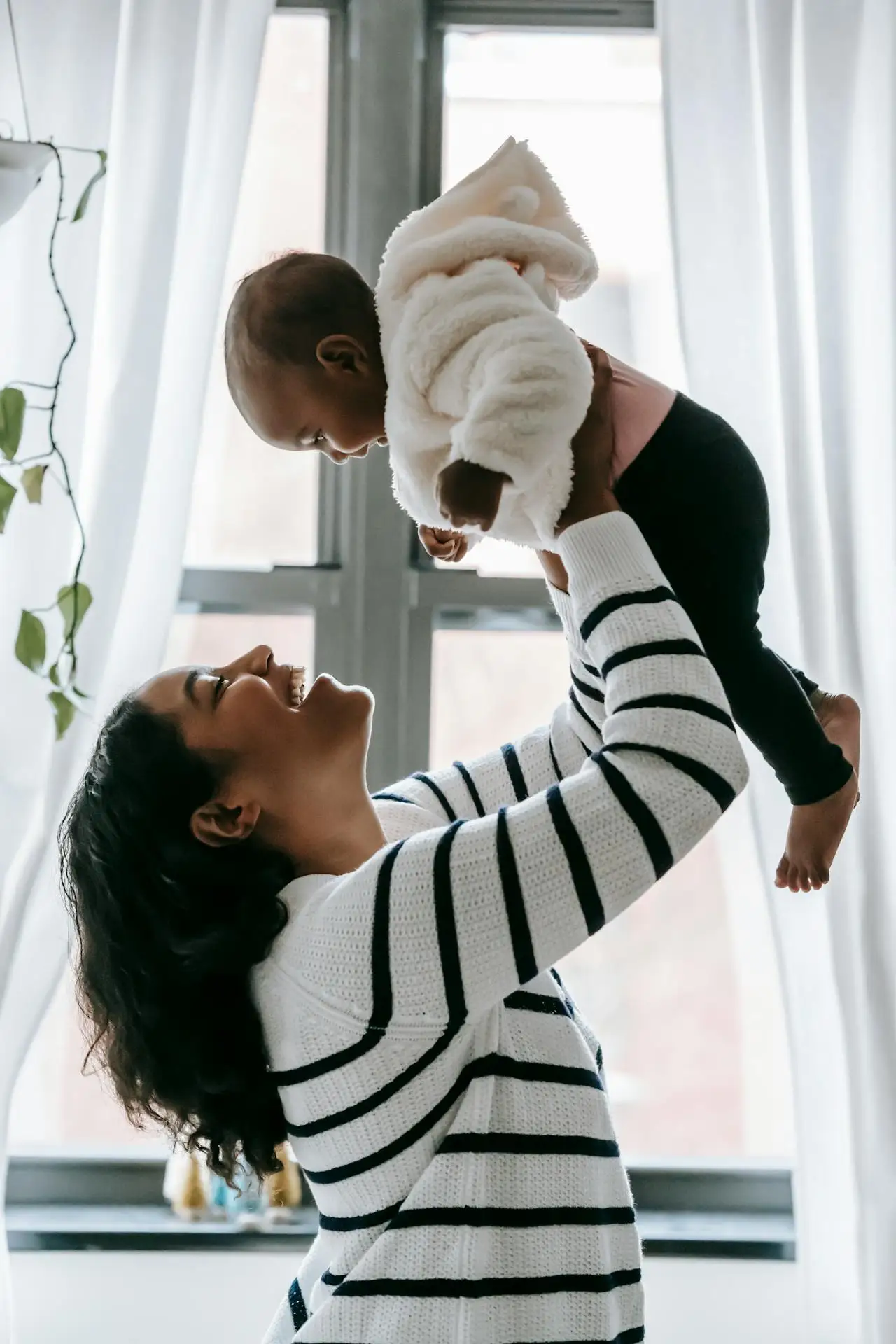 Mãe e filho interagindo em casa: a criança usa pijama branco com listras pretas e cobertor de pelúcia, enquanto a mãe o abraça em um ambiente iluminado pela janela.