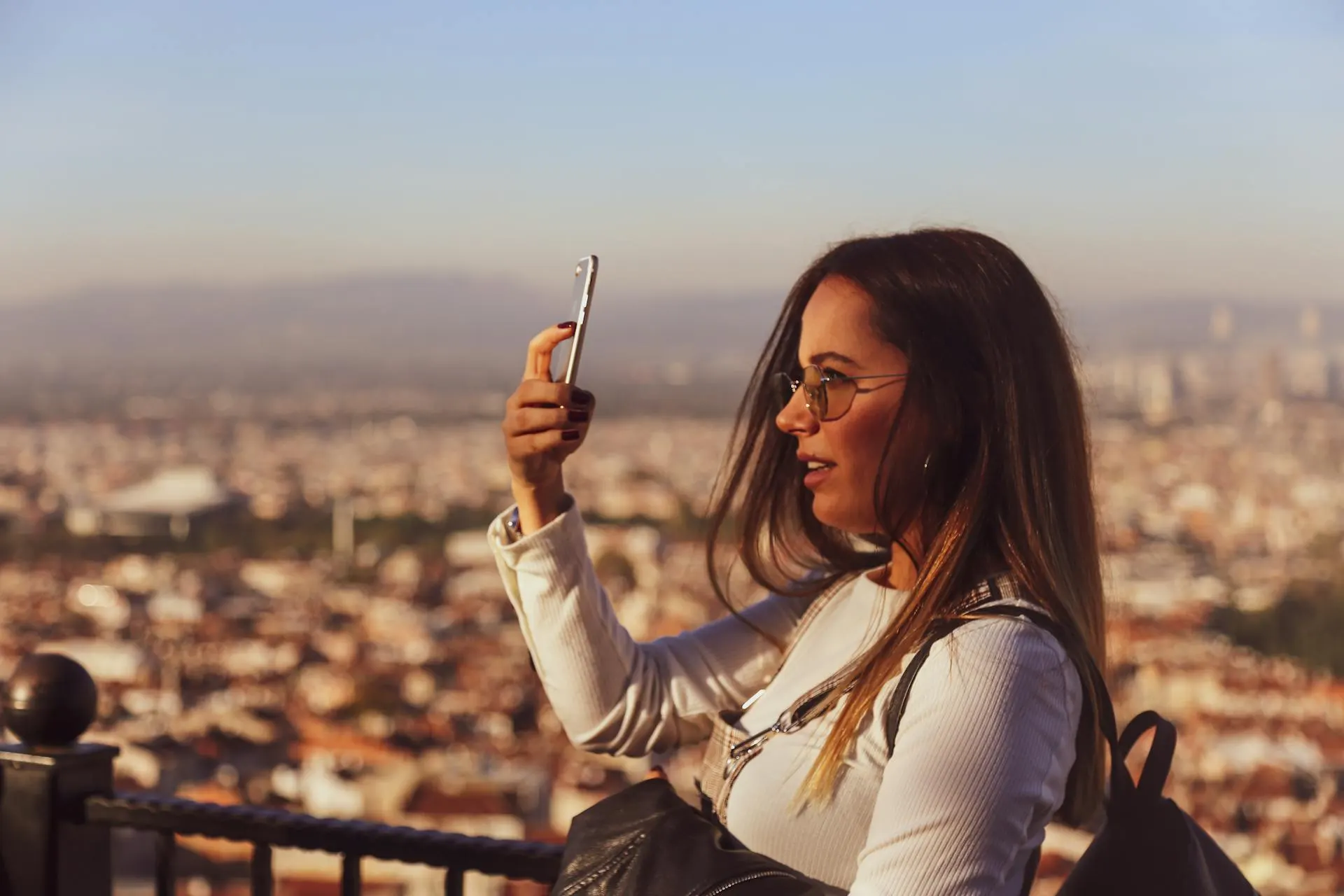 Mulher jovem com óculos e cabelo longo usa uma jaqueta clara e segura um celular na mão, sentada em um mirante com vista panorâmica da cidade ao entardecer, usando mochila.